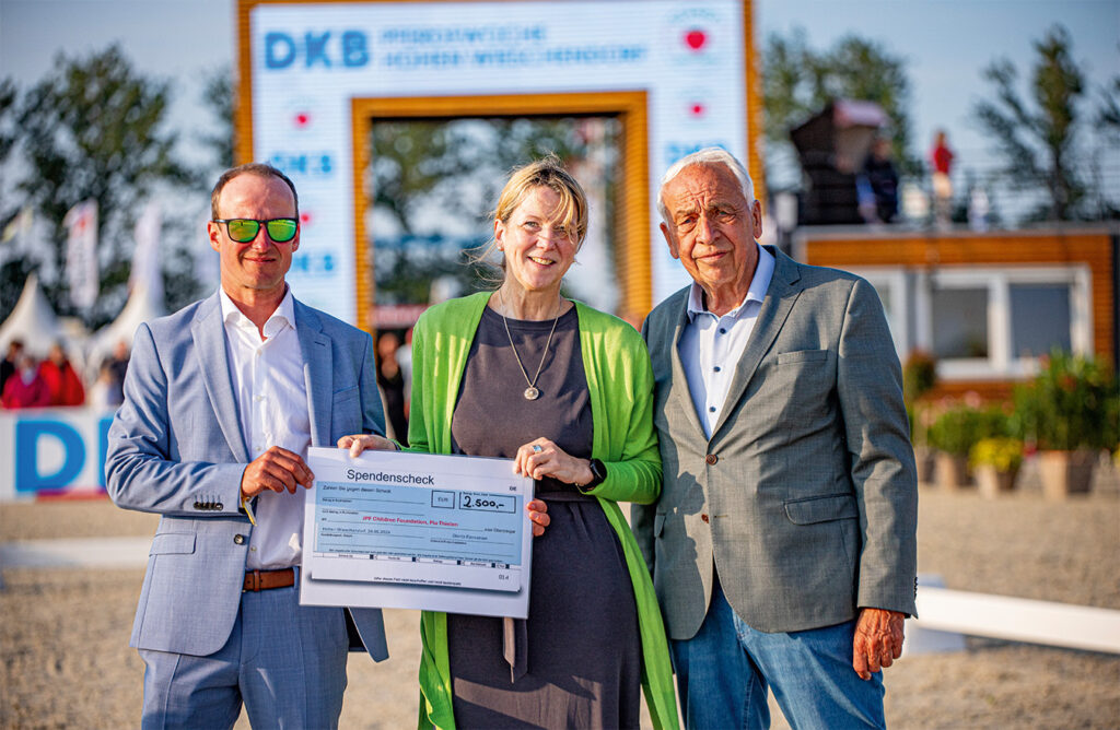 Drei Personen lächeln und halten einen Scheck im Freien, ein Mann trägt Sonnenbrille.