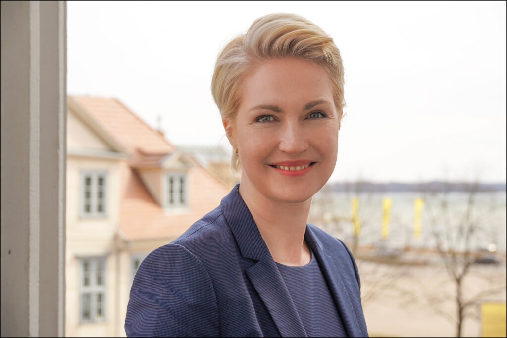 Eine lächelnde Person mit blonder Kurzhaarfrisur in einem blauen Blazer vor einem Haus.