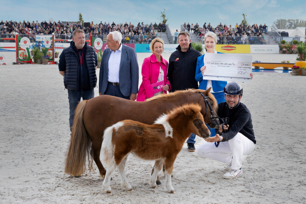 Eine Gruppe freut sich neben Ponys; Person kniet mit Helm; Frau hält Scheck; helle Outfits, sandiger Boden.